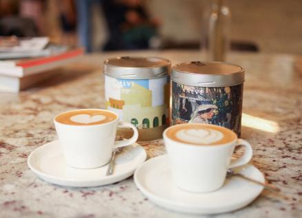Divas kafijas tasītes ar sirds formas latte mākslu uz marmora galda. Aiz tām redzamas divas "Kalve" kafijas bundžas ar mākslinieciskiem dizainiem un uzrakstiem "Espresso blend of arts" un "Signature espresso blend. Toffee". Fonā – grāmatu kaudzīte un izpludināts interjera fons.