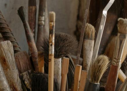 Dažāda izmēra otu un instrumentu saišķis ar nolietotiem rokturiem, cieši salikti kopā putekļainā telpā.
