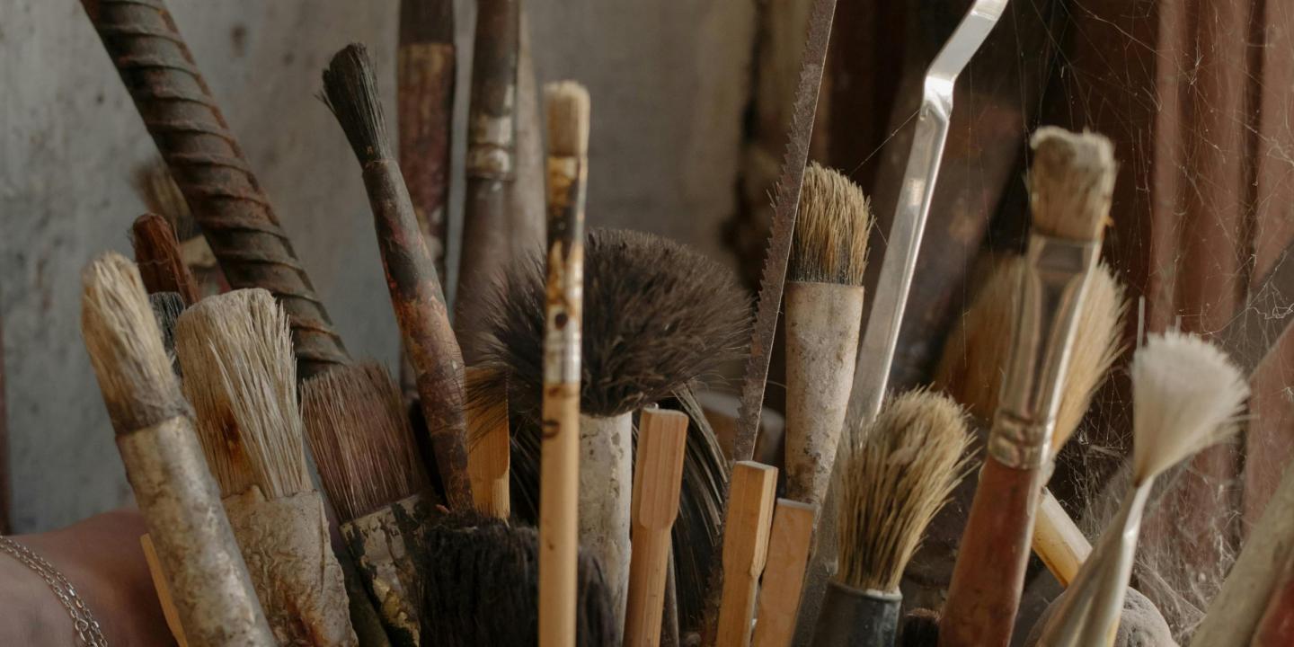 Dažāda izmēra otu un instrumentu saišķis ar nolietotiem rokturiem, cieši salikti kopā putekļainā telpā.