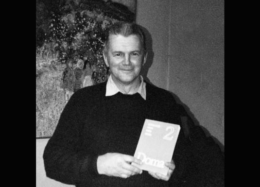 Melnbalta fotogrāfija, kurā redzams vīrietis, stāvot telpā pie gleznas, rokās turot grāmatu vai katalogu.