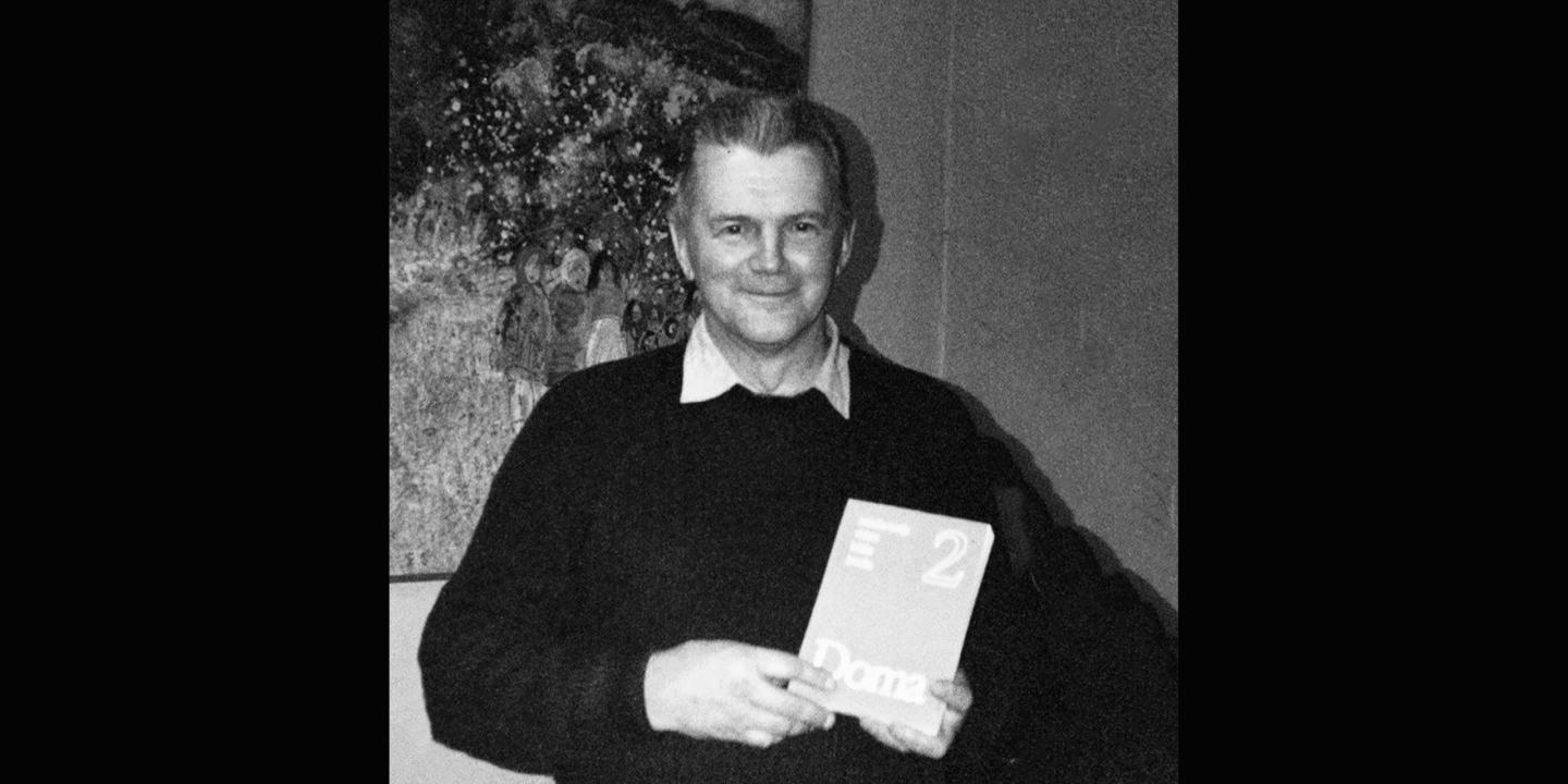 Melnbalta fotogrāfija, kurā redzams vīrietis, stāvot telpā pie gleznas, rokās turot grāmatu vai katalogu.