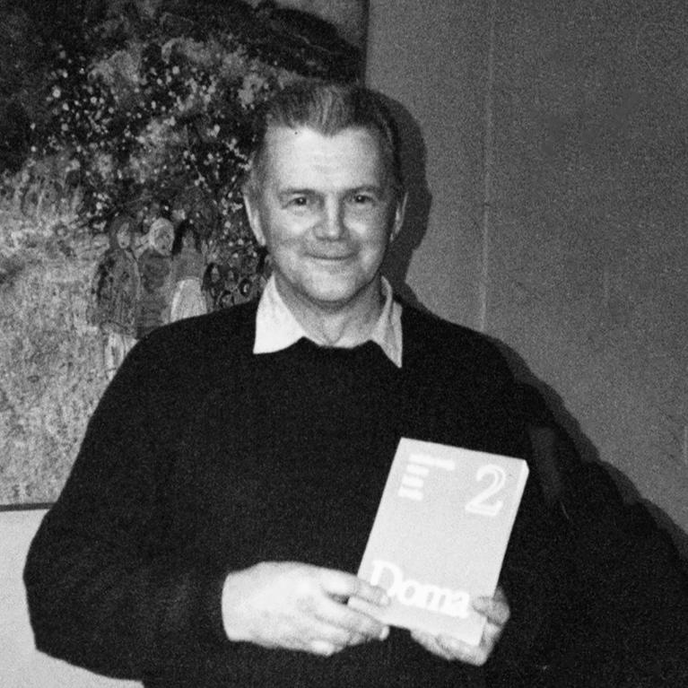 Melnbalta fotogrāfija, kurā redzams vīrietis, stāvot telpā pie gleznas, rokās turot grāmatu vai katalogu.