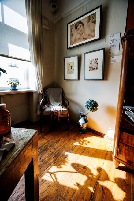 Gaiša interjera telpas stūrī pie loga novietots krēsls ar segu, pie sienas redzami ierāmēti zīmējumi, uz grīdas vāze ar ziedu.