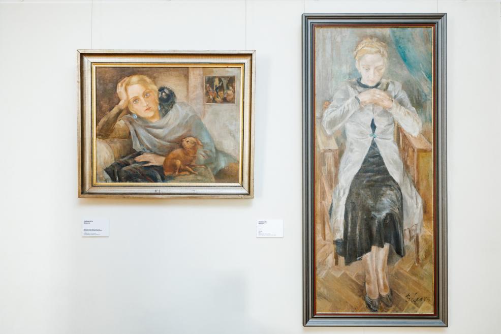 Pie sienas izstādītas divas gleznas: vienā attēlota sieviete ar suni, otrā – sēdoša sieviete gaišā jakā.