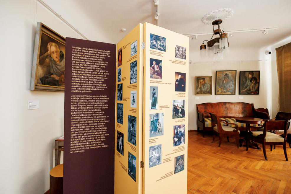 Muzeja telpā redzams informatīvs stends ar attēliem un tekstiem, fonā izvietotas gleznas un vēsturiskas mēbeles ar dīvānu un krēsliem.