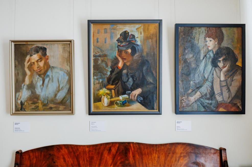 Pie sienas izvietotas trīs gleznas ar portretiem, zem tām redzama koka sola atzveltne.