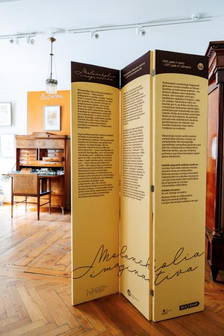 Muzeja telpā izvietots salokāms informatīvs stends ar tekstiem par izstādi “Melanholija imaginatīva”, fonā redzamas vēsturiskas mēbeles.