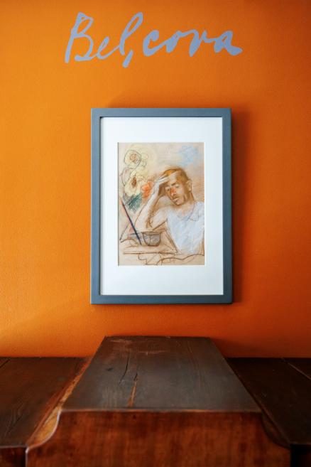 Uz oranžas sienas ar uzrakstu “Belcova” izvietots ierāmēts zīmējums ar sievietes portretu, zem tā redzama koka virsma.