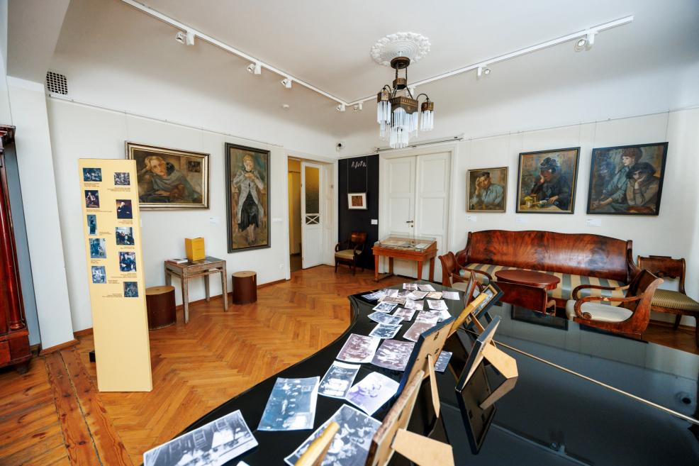 Muzeja telpā ar parketa grīdu redzama ekspozīcija ar gleznām pie sienām, koka mēbelēm un galdu ar izliktām fotogrāfijām.