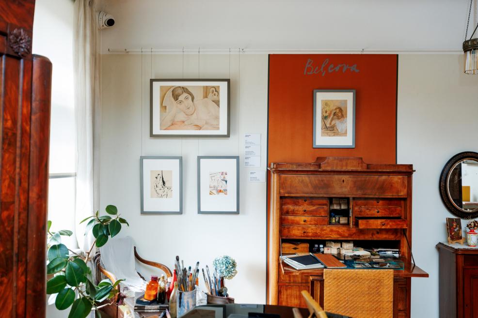 Muzeja ekspozīcijā redzams interjers ar koka rakstāmgaldu, zīmējumiem pie sienas, oranžu paneli ar uzrakstu “Belcova” un priekšplānā mākslinieka darbarīkiem.