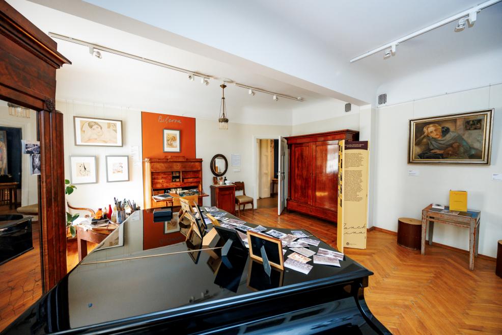 Plaša muzeja telpa ar parketa grīdu, kur redzams klavieru vāks ar fotogrāfijām, koka mēbeles, gleznas pie sienām un oranžs “Belcova” ekspozīcijas panelis.