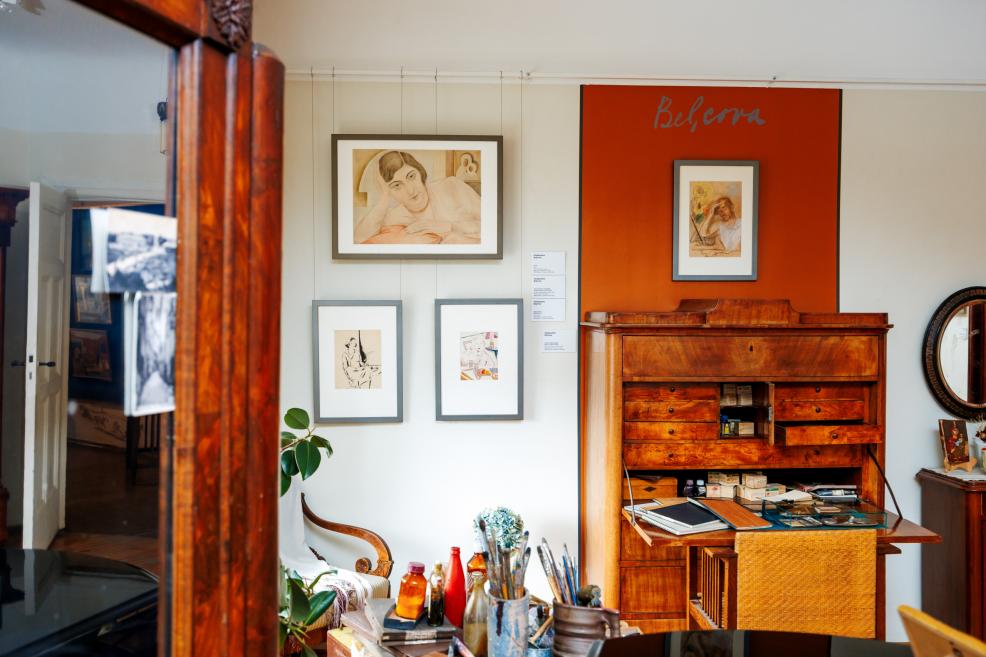 Muzeja ekspozīcijā redzams koka rakstāmgalds ar atvilktnēm, pie sienas izkārti zīmējumi un oranžs “Belcova” panelis, priekšplānā mākslinieka darbarīki.