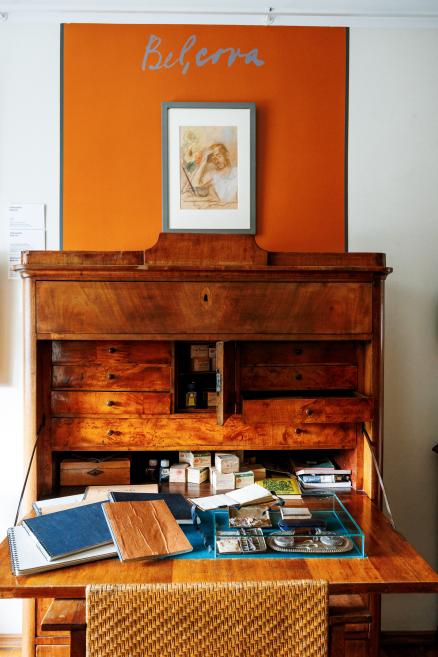 Koka rakstāmgalds ar atvērtām atvilktnēm, uz kura izlikti pieraksti un priekšmeti, virs tā oranžs panelis ar uzrakstu “Belcova” un ierāmēts zīmējums.