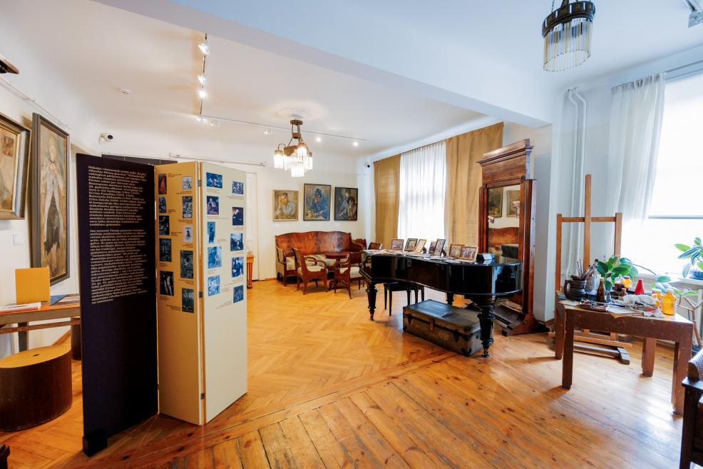 Plaša muzeja telpa ar koka grīdu, kur redzamas gleznas pie sienām, informatīvs stends, klavieres ar fotogrāfijām un mākslinieka darbnīcas galds pie loga.