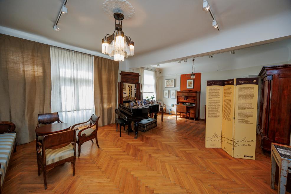 Plaša muzeja telpa ar parketa grīdu, kur redzamas klavieres, koka mēbeles, informatīvs stends un gleznas pie sienām.