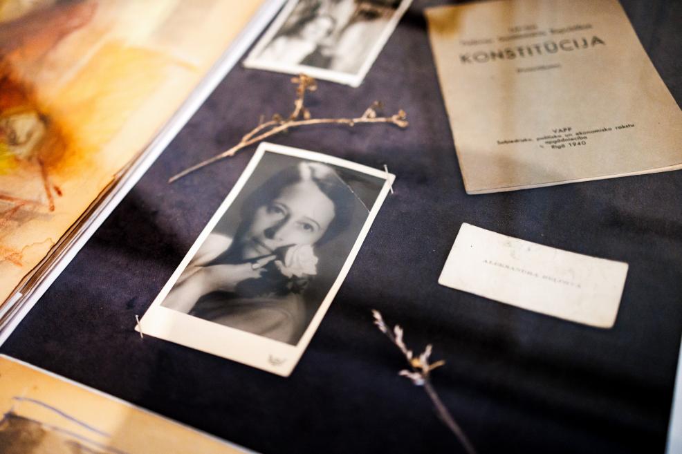  Uz galda izkārtotas melnbaltas fotogrāfijas, žāvēti augu zariņi un dokuments ar uzrakstu “Konstitūcija”.