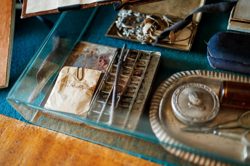 Stikla vitrīnā redzami zīmēšanas rīki, krāsu palete, papīrs ar rokrakstu un nelieli personiski priekšmeti.