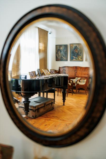 Spogulī redzams muzeja interjers ar klavierēm, fotogrāfijām un gleznām pie sienas.