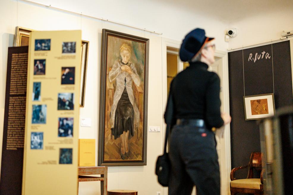 Muzeja telpā apmeklētāja skatās gleznas, fonā redzama ekspozīcija ar darbiem un informatīvu stendu.