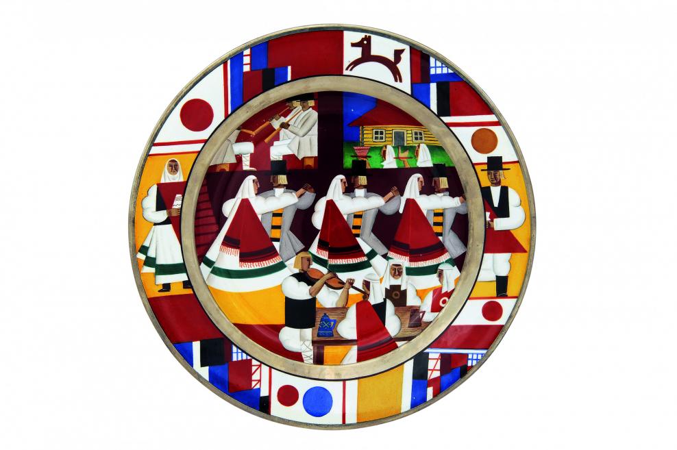Dekoratīvs apaļš šķīvis ar stilizētu folkloras ainu, kurā attēloti tautastērpos ģērbti cilvēki dejas un rituāla kustībās, ģeometrisku rakstu un košu krāsu kompozīcijā.