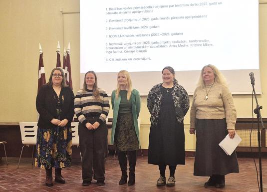 Attēlā redzamas piecas sievietes rindā zāles priekšā, fonā redzami Latvijas karogi un ar projektoru izgaismota balta fona siena, kur attēlots teksts ar plāna punktiem.