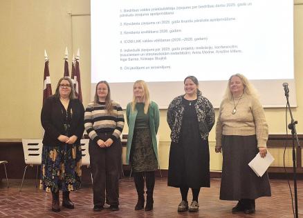 Attēlā redzamas piecas sievietes rindā zāles priekšā, fonā redzami Latvijas karogi un ar projektoru izgaismota balta fona siena, kur attēlots teksts ar plāna punktiem.