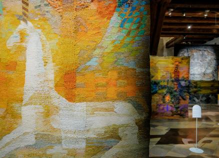 Muzeja ekspozīcijas skats ar lielizmēra tekstilmākslas darbiem siltos dzeltenos, oranžos un zaļos toņos; priekšplānā abstrakts auduma gobelēns, fonā vairāki piekārti tekstila objekti vēsturiskā telpā.