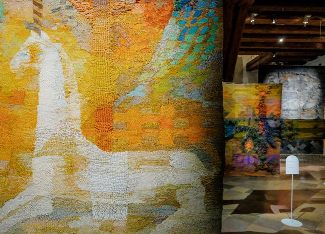 Muzeja ekspozīcijas skats ar lielizmēra tekstilmākslas darbiem siltos dzeltenos, oranžos un zaļos toņos; priekšplānā abstrakts auduma gobelēns, fonā vairāki piekārti tekstila objekti vēsturiskā telpā.