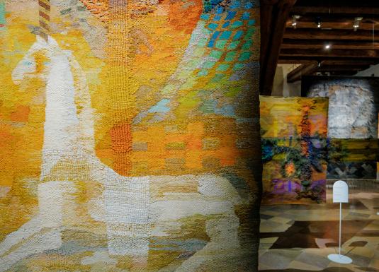 Muzeja ekspozīcijas skats ar lielizmēra tekstilmākslas darbiem siltos dzeltenos, oranžos un zaļos toņos; priekšplānā abstrakts auduma gobelēns, fonā vairāki piekārti tekstila objekti vēsturiskā telpā.