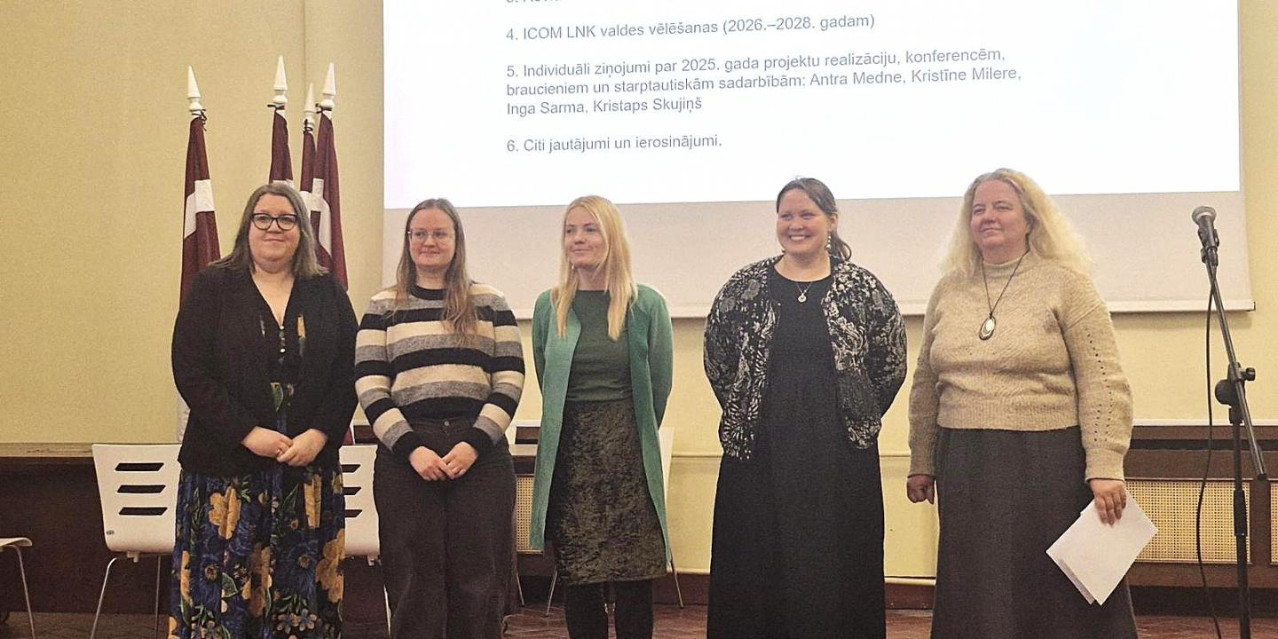 Attēlā redzamas piecas sievietes rindā zāles priekšā, fonā redzami Latvijas karogi un ar projektoru izgaismota balta fona siena, kur attēlots teksts ar plāna punktiem.