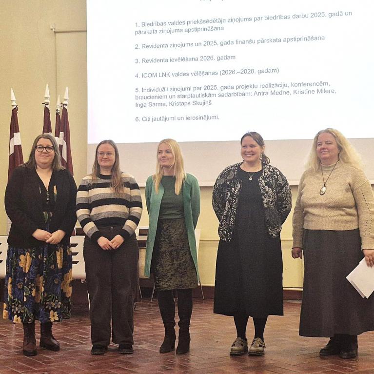 Attēlā redzamas piecas sievietes rindā zāles priekšā, fonā redzami Latvijas karogi un ar projektoru izgaismota balta fona siena, kur attēlots teksts ar plāna punktiem.