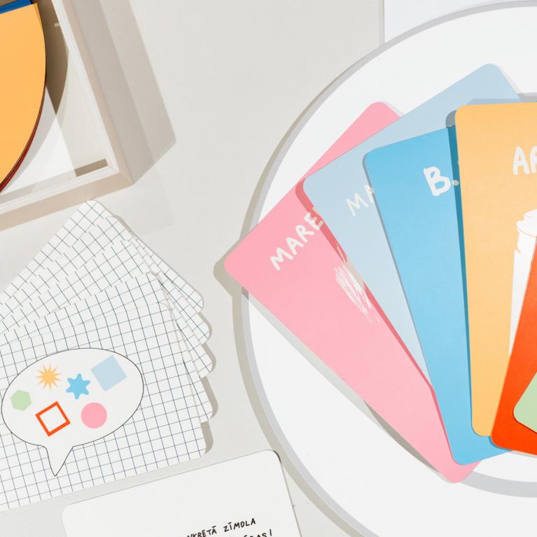 Izstādes dizaina elementi – krāsainas kartītes, drukāti materiāli un grafiski objekti, izkārtoti uz balta galda.