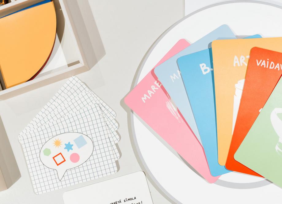 Izstādes dizaina elementi – krāsainas kartītes, drukāti materiāli un grafiski objekti, izkārtoti uz balta galda.