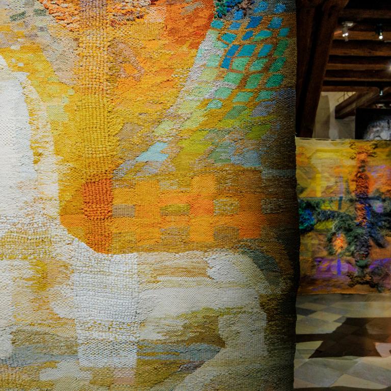 Muzeja ekspozīcijas skats ar lielizmēra tekstilmākslas darbiem siltos dzeltenos, oranžos un zaļos toņos; priekšplānā abstrakts auduma gobelēns, fonā vairāki piekārti tekstila objekti vēsturiskā telpā.