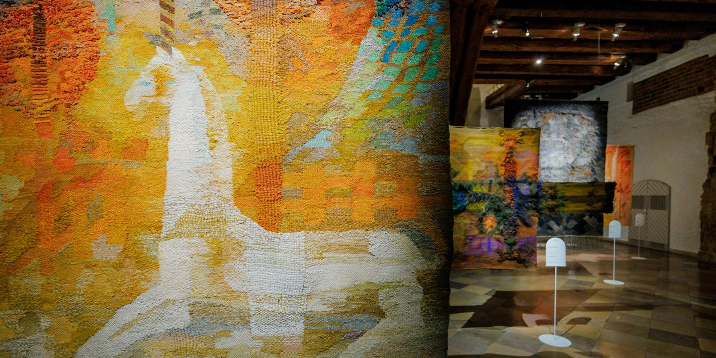 Muzeja ekspozīcijas skats ar lielizmēra tekstilmākslas darbiem siltos dzeltenos, oranžos un zaļos toņos; priekšplānā abstrakts auduma gobelēns, fonā vairāki piekārti tekstila objekti vēsturiskā telpā.
