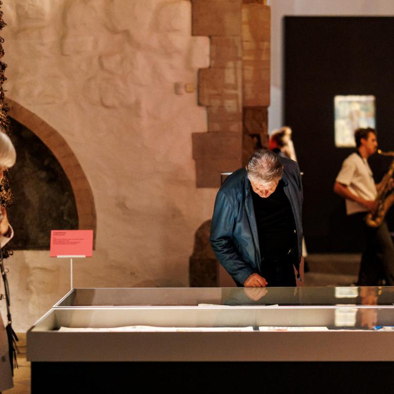 Muzeja izstāžu zālē divi apmeklētāji aplūko stiklotu vitrīnu ar ekspozīciju, fonā redzami mākslas darbi un saksafonists.