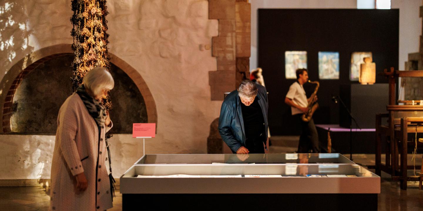 Muzeja izstāžu zālē divi apmeklētāji aplūko stiklotu vitrīnu ar ekspozīciju, fonā redzami mākslas darbi un saksafonists.