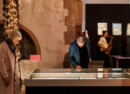 Muzeja izstāžu zālē divi apmeklētāji aplūko stiklotu vitrīnu ar ekspozīciju, fonā redzami mākslas darbi un saksafonists.