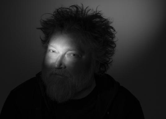 Tumšā portretā redzams vīrietis ar izspūrušiem matiem un bārdu, kura seja daļēji izgaismota ar koncentrētu gaismas staru.