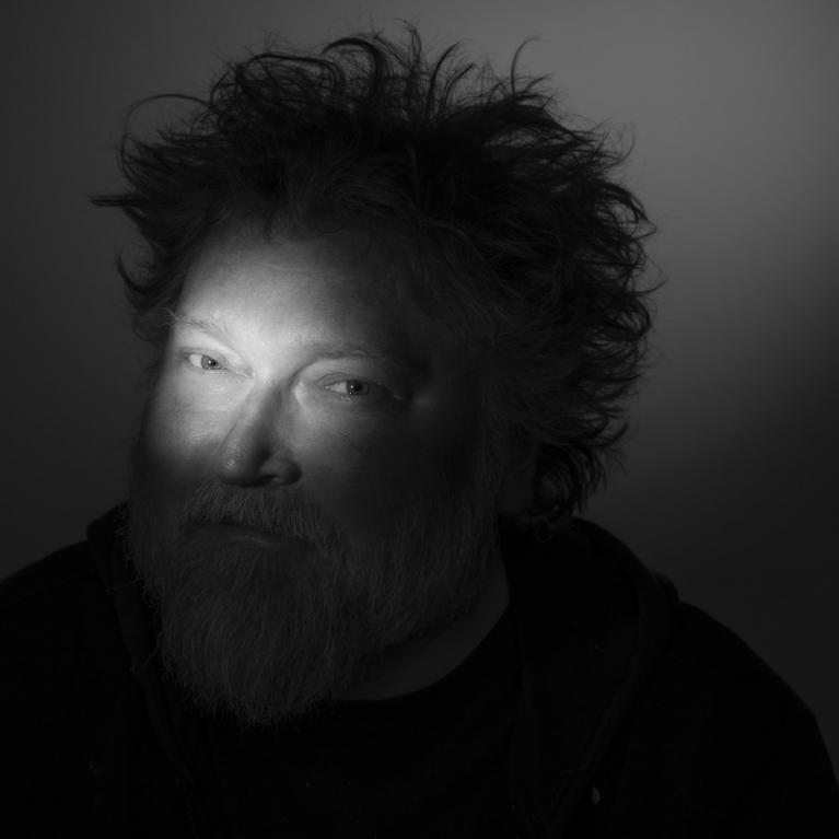 Tumšā portretā redzams vīrietis ar izspūrušiem matiem un bārdu, kura seja daļēji izgaismota ar koncentrētu gaismas staru.