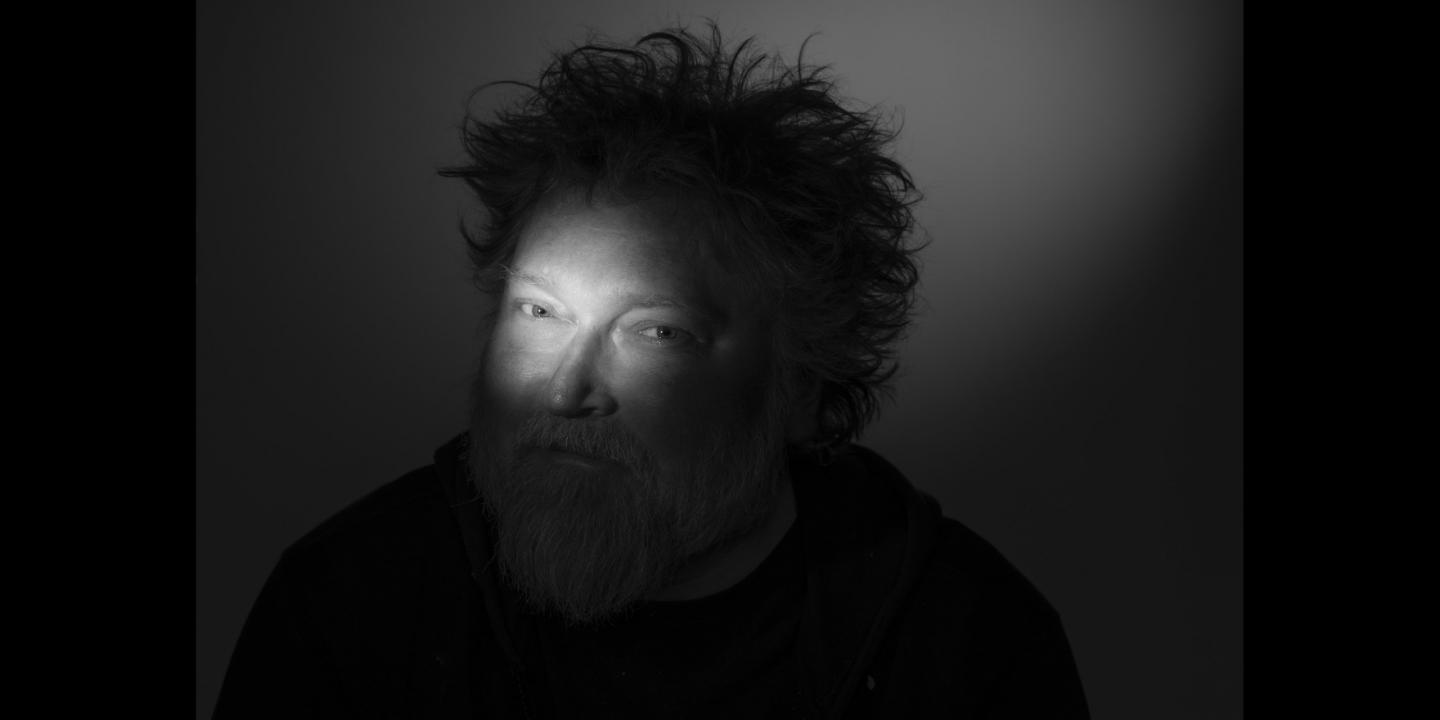 Tumšā portretā redzams vīrietis ar izspūrušiem matiem un bārdu, kura seja daļēji izgaismota ar koncentrētu gaismas staru.