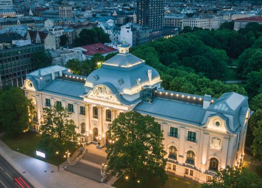 Apgaismota Latvijas Nacionālā mākslas muzeja ēka Rīgā krēslā, skatīta no augšas ar pilsētas panorāmu fonā.