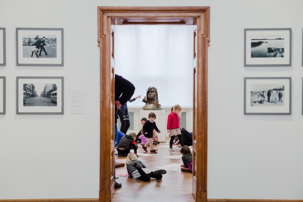 Bērni muzeja telpā sēž un spēlējas uz grīdas, fonā redzama skulptūra un pie sienām izvietotas fotogrāfijas.