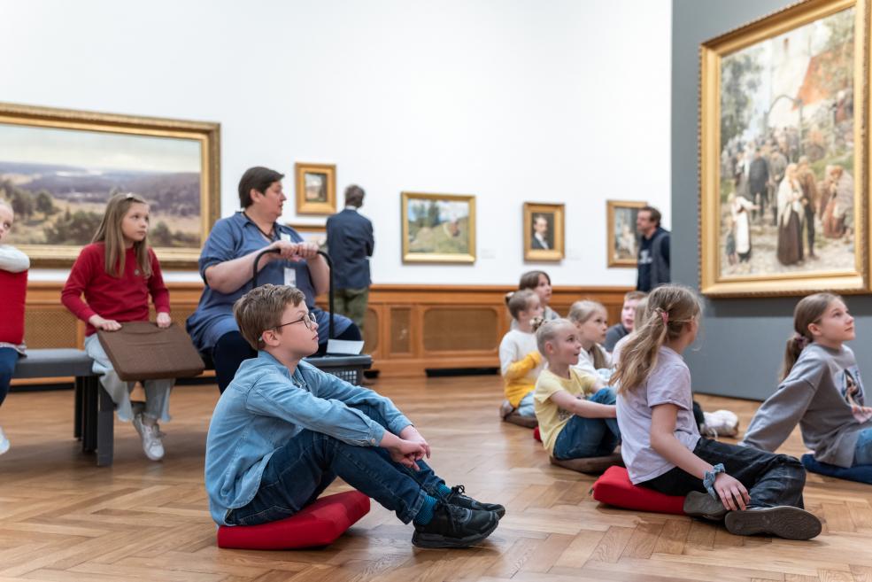 Bērni muzeja zālē sēž uz spilveniem un klausās, kamēr pie sienām redzamas gleznas un pieaugušais viņus uzrauga.