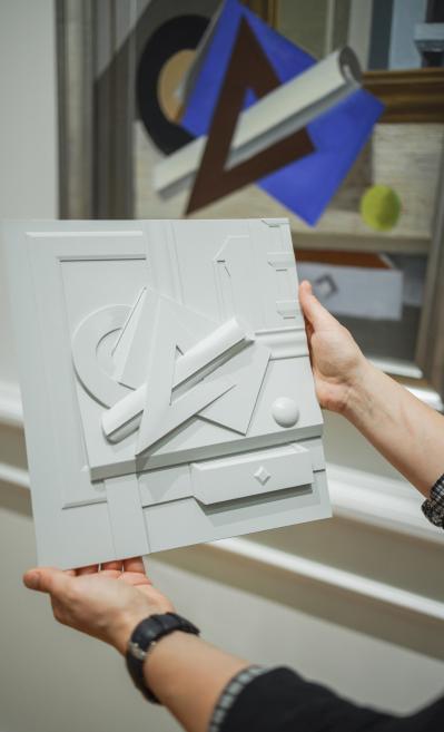 Rokās turēts reljefs balts mākslas objekts ar ģeometriskām formām, fonā redzama krāsaina abstrakta kompozīcija.