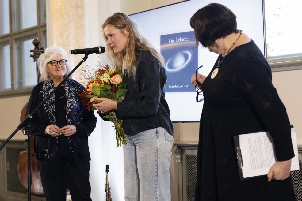 Sieviete pasniedz runu pie mikrofona, rokās turot ziedus, kamēr blakus stāv divas citas sievietes un fonā redzams prezentācijas ekrāns.