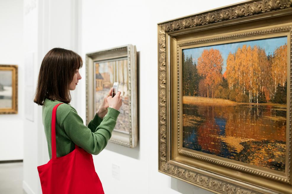 Sieviete muzejā fotografē rudens ainavas gleznu ar telefonu.