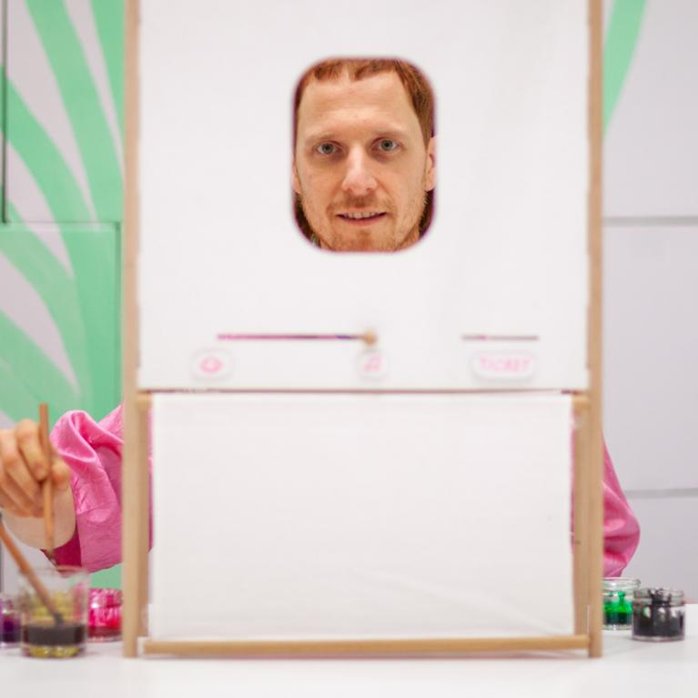 Man behind a small frame with a face opening, painting with brushes and paints on a table.