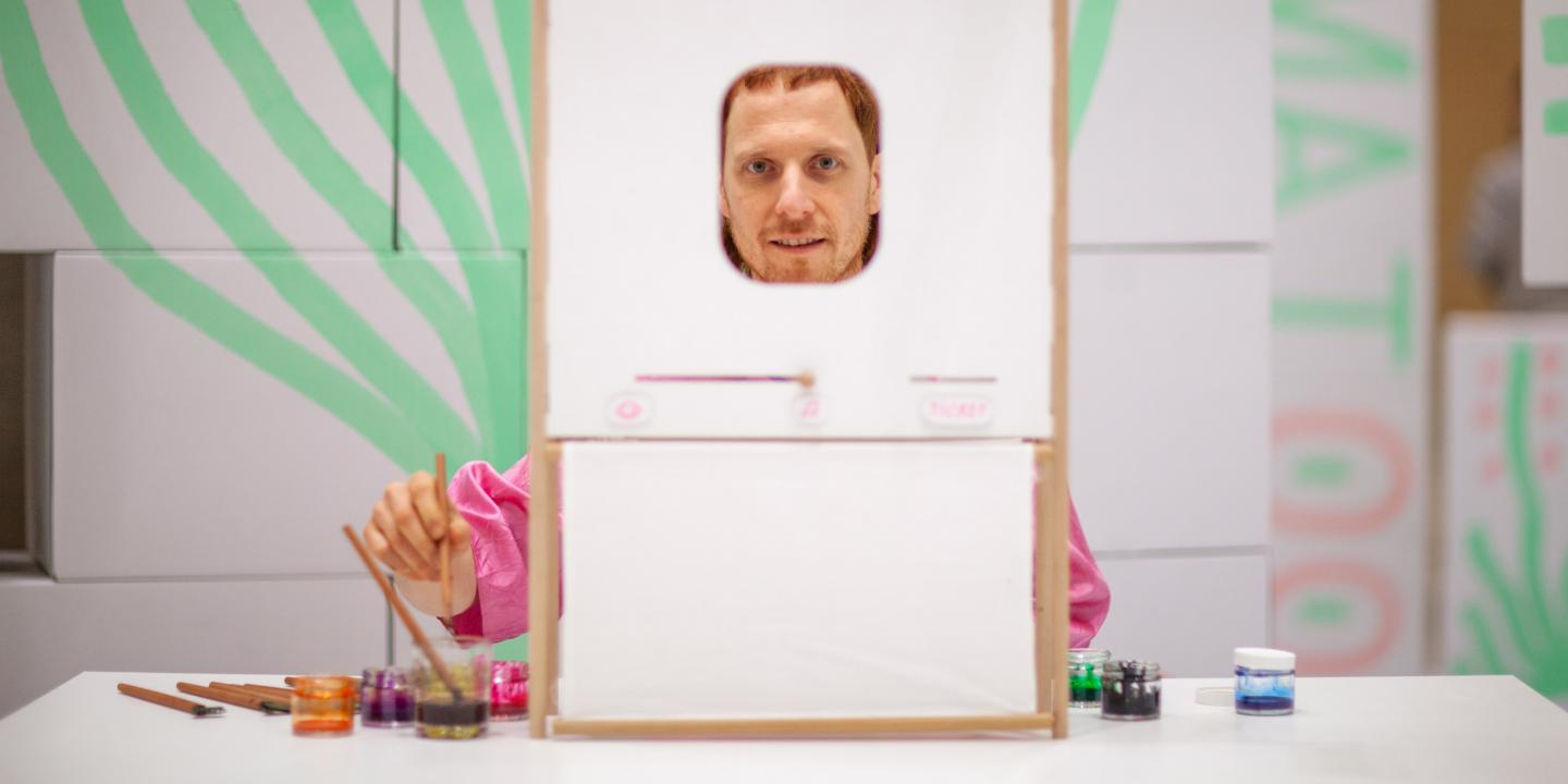 Man behind a small frame with a face opening, painting with brushes and paints on a table.