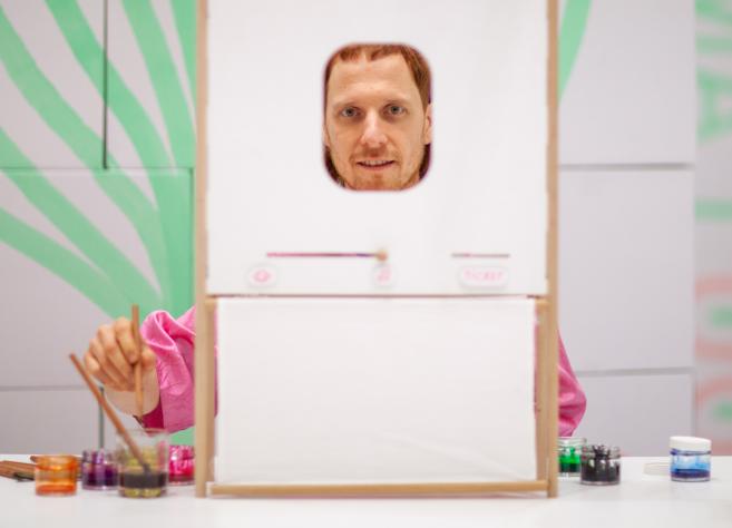 Man behind a small frame with a face opening, painting with brushes and paints on a table.